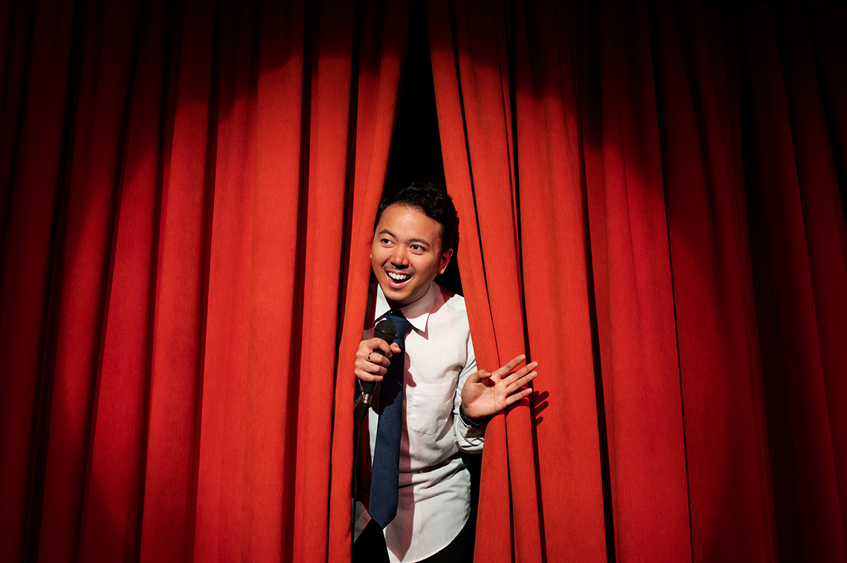 Image of a comedian peeking through the curtains on a stage holding a microphone